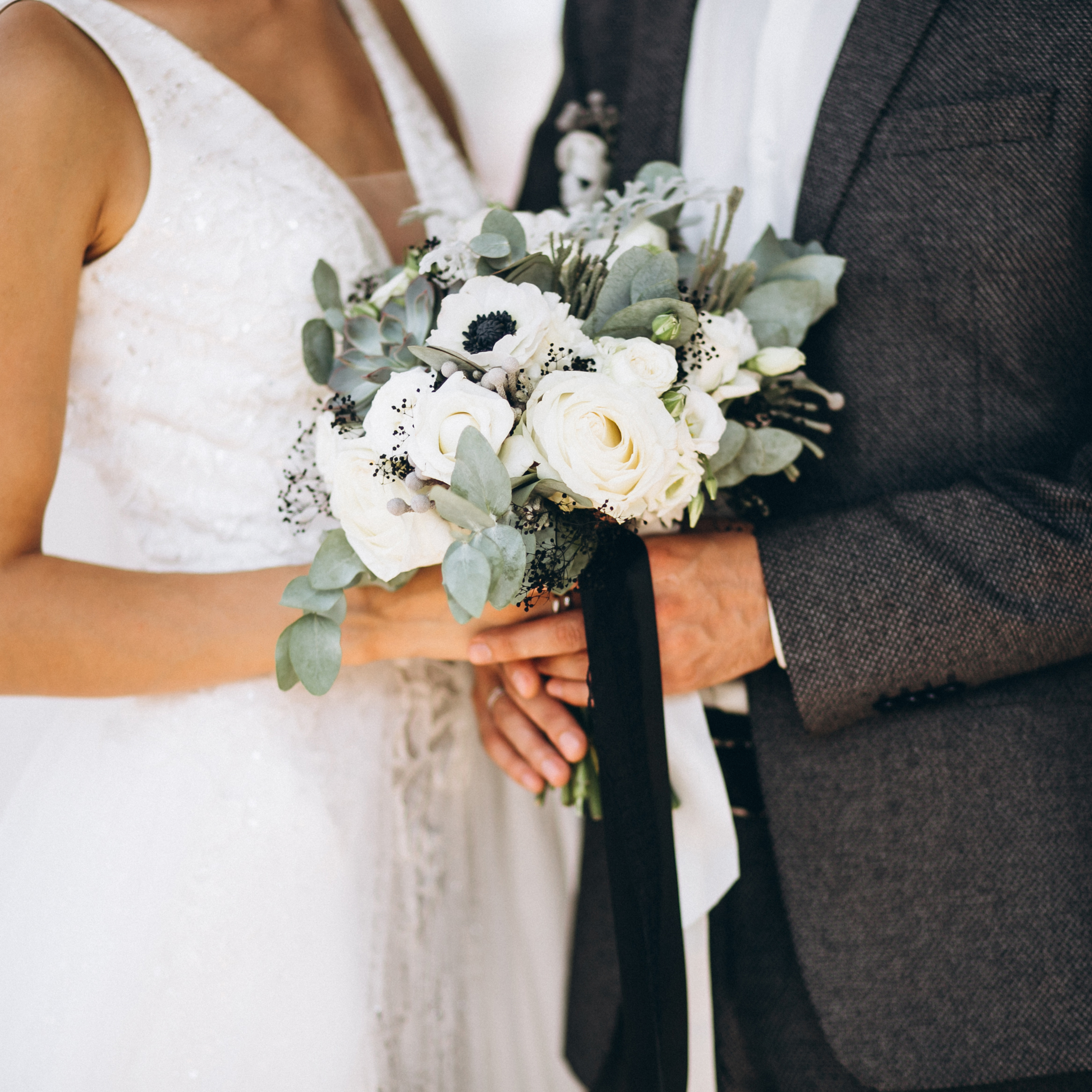 Ein Paar hält gemeinsam einen Brautstrauß aus weißen Rosen, umgeben von grünem Laub, bei einer Hochzeit.