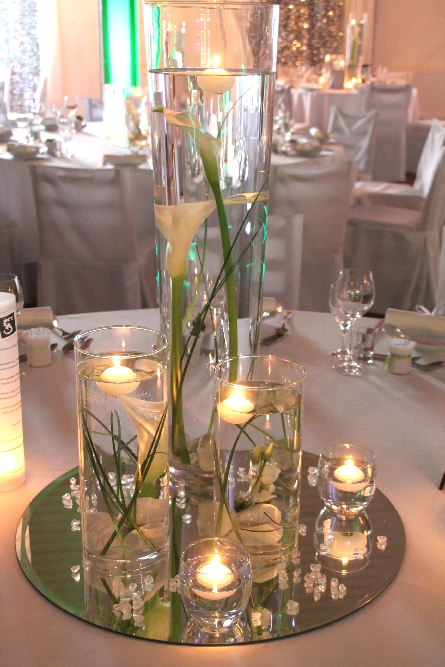 Elegante Tischdekoration mit Glasvasen, weißen Calla-Lilien und schwimmenden Kerzen auf einem Spiegel.