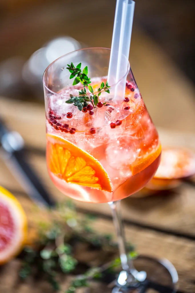 Ein elegantes Weinglas mit einem orange-rosa Cocktail, verziert mit Orangenscheiben, Beeren und Thymian.